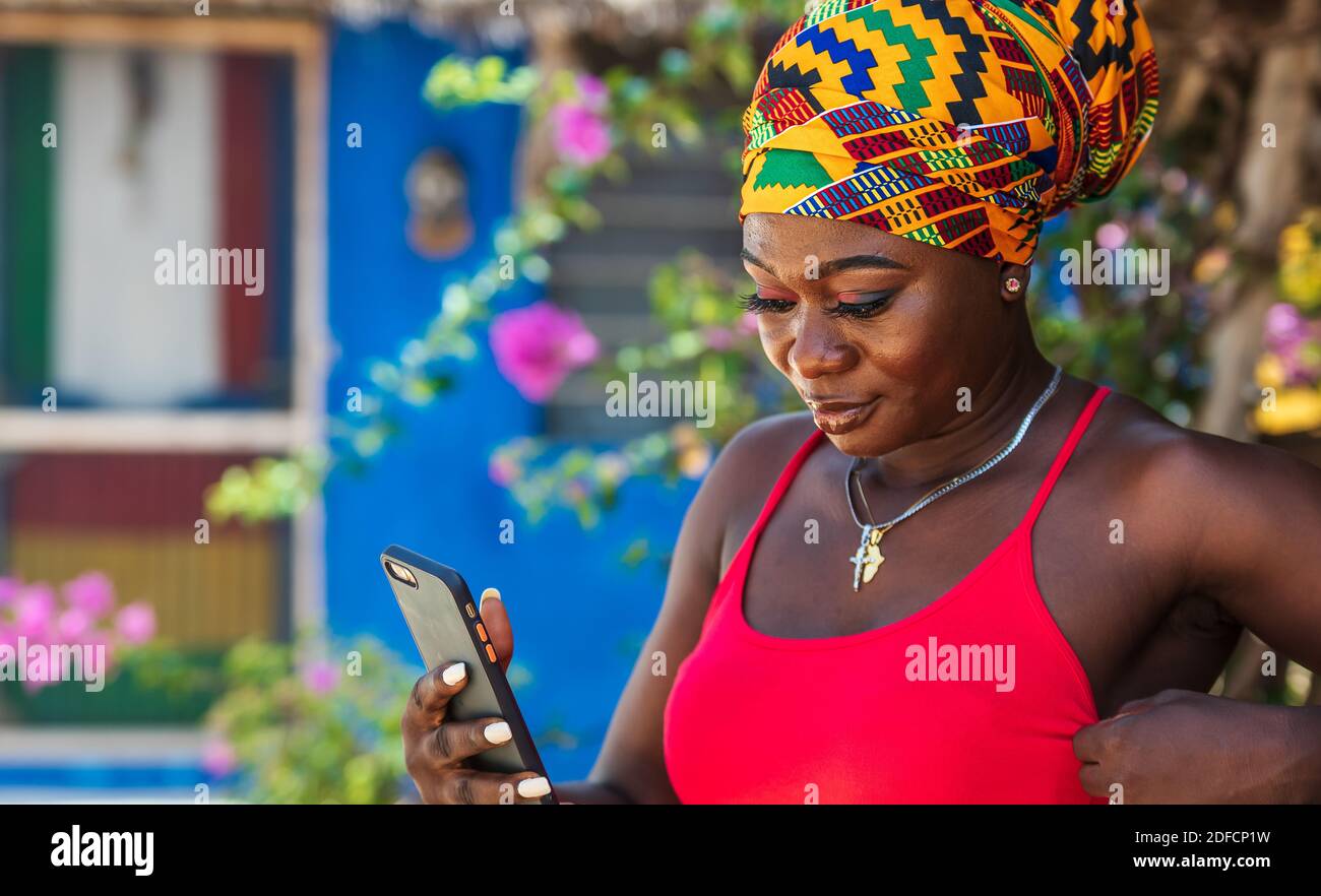 Afrikanische Frau mit afrikanischem Kopfschmuck auf dem tropischen Teil von Ghana mit Handy in der Hand Stockfoto
