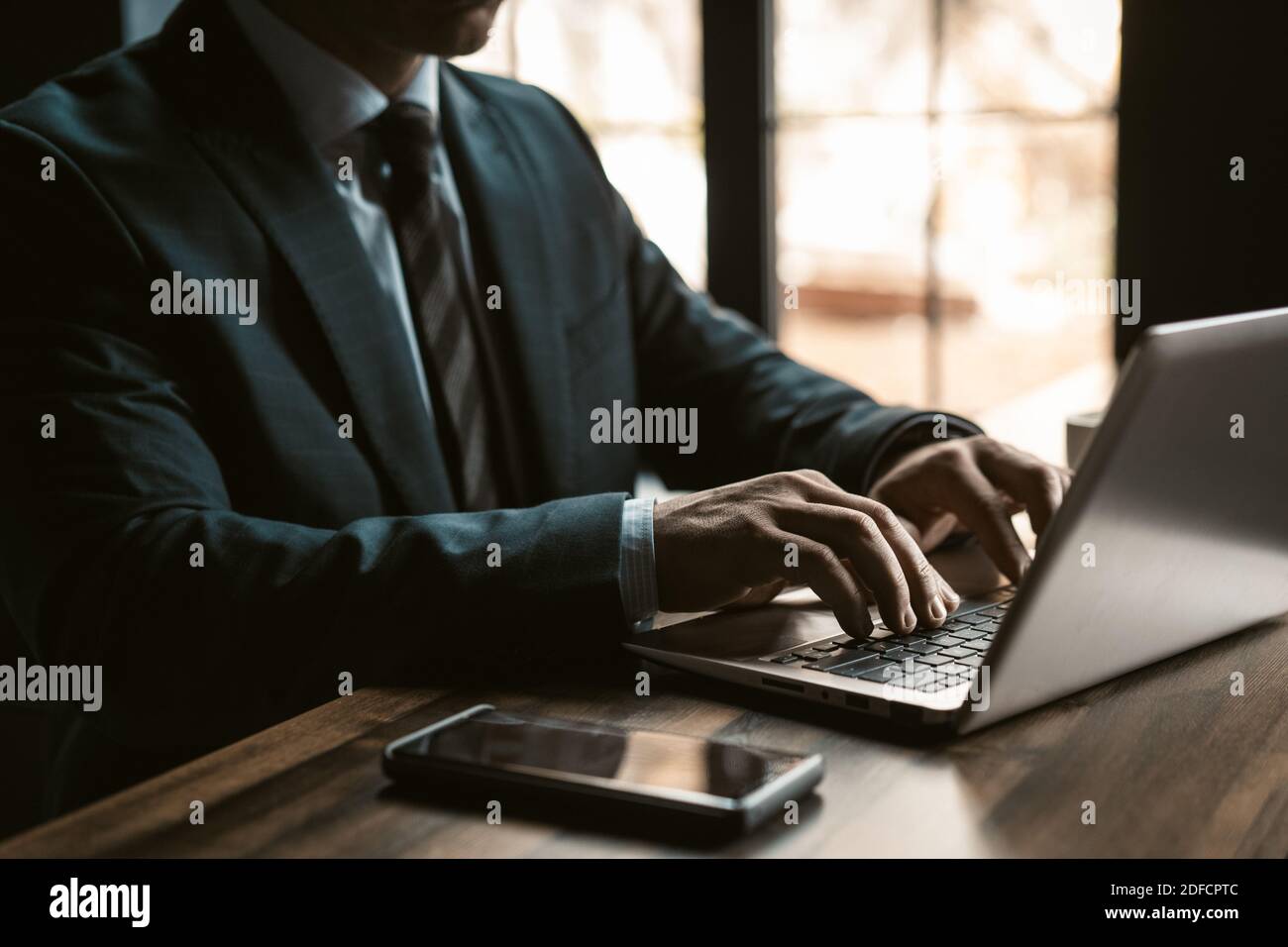 Nahaufnahme. Geschäftsmann in einer teuren Business-Suite, die in einem modernen Büro mit einem Laptop und einem Smartphone daneben arbeitet und Fenster im Hintergrund hat. Mann, der arbeitet Stockfoto