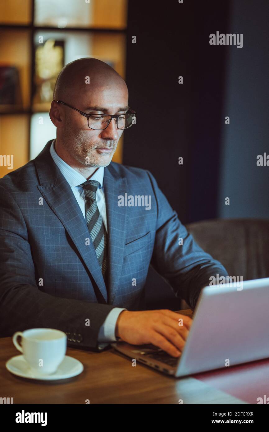 Mittelalter, kahle Geschäftsmann in der Suite, der an einem Laptop im modernen Büro sitzt. Gut aussehend kühnen Mann arbeitet am Computer im Büro mit einer Tasse Stockfoto