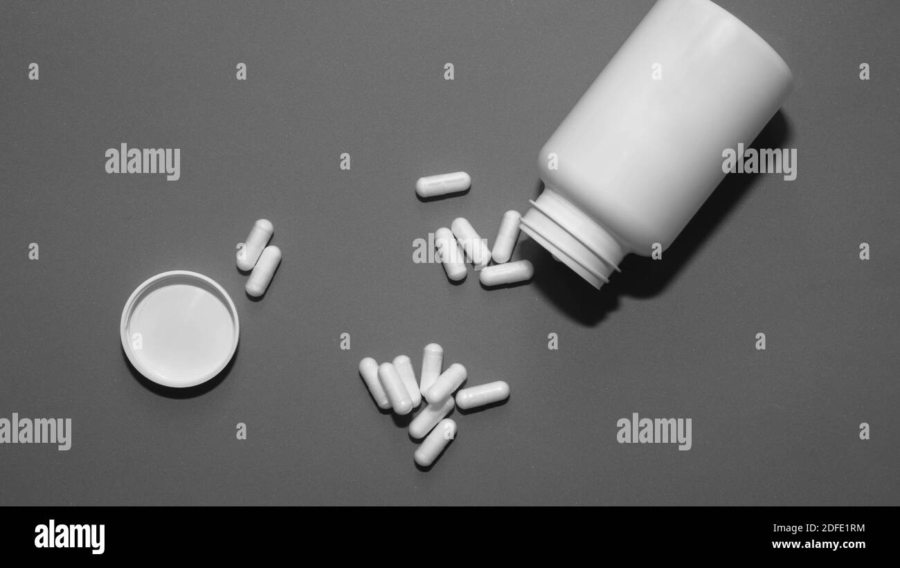 Weißes leeres medizinisches Glas, verstreute Tabletten in Kapseln, auf grauem Hintergrund. Stockfoto