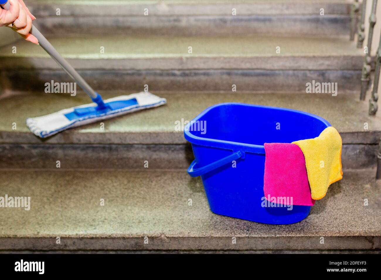 Reinigungsgeräte, ein blauer Kunststoffeimer mit gelbem und rosa Mikrofasertuch, auf grauen Granittreppen stehend. Eine Hand, die den Wischmopp hält, der die Treppe abwischt Stockfoto
