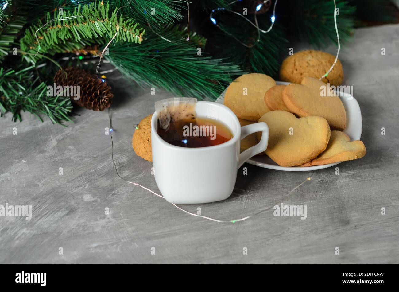 Hausgemachte Kekse in Form eines Herzens auf einem weißen Teller mit einem Becher und einem Teebeutel auf dem Hintergrund der Weihnachtsdekoration. Selektiver Fokus. Stockfoto