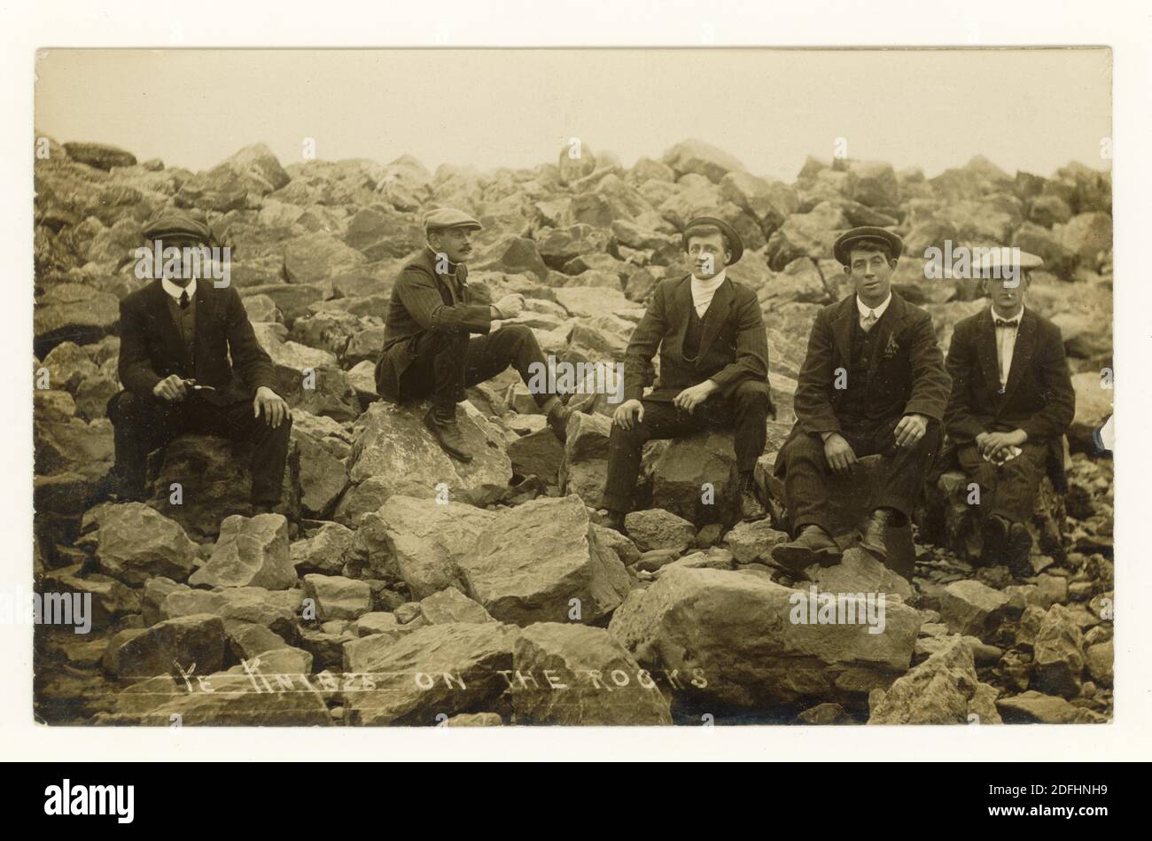 Postkarte der frühen 1900er Jahre von jungen Männern in flachen Kappen und tragen ihre 'Sunday Best' Kleidung, die auf Felsen sitzt - seine Knibs geschrieben auf der Vorderseite - circa Anfang der 1920er Jahre, Großbritannien Stockfoto