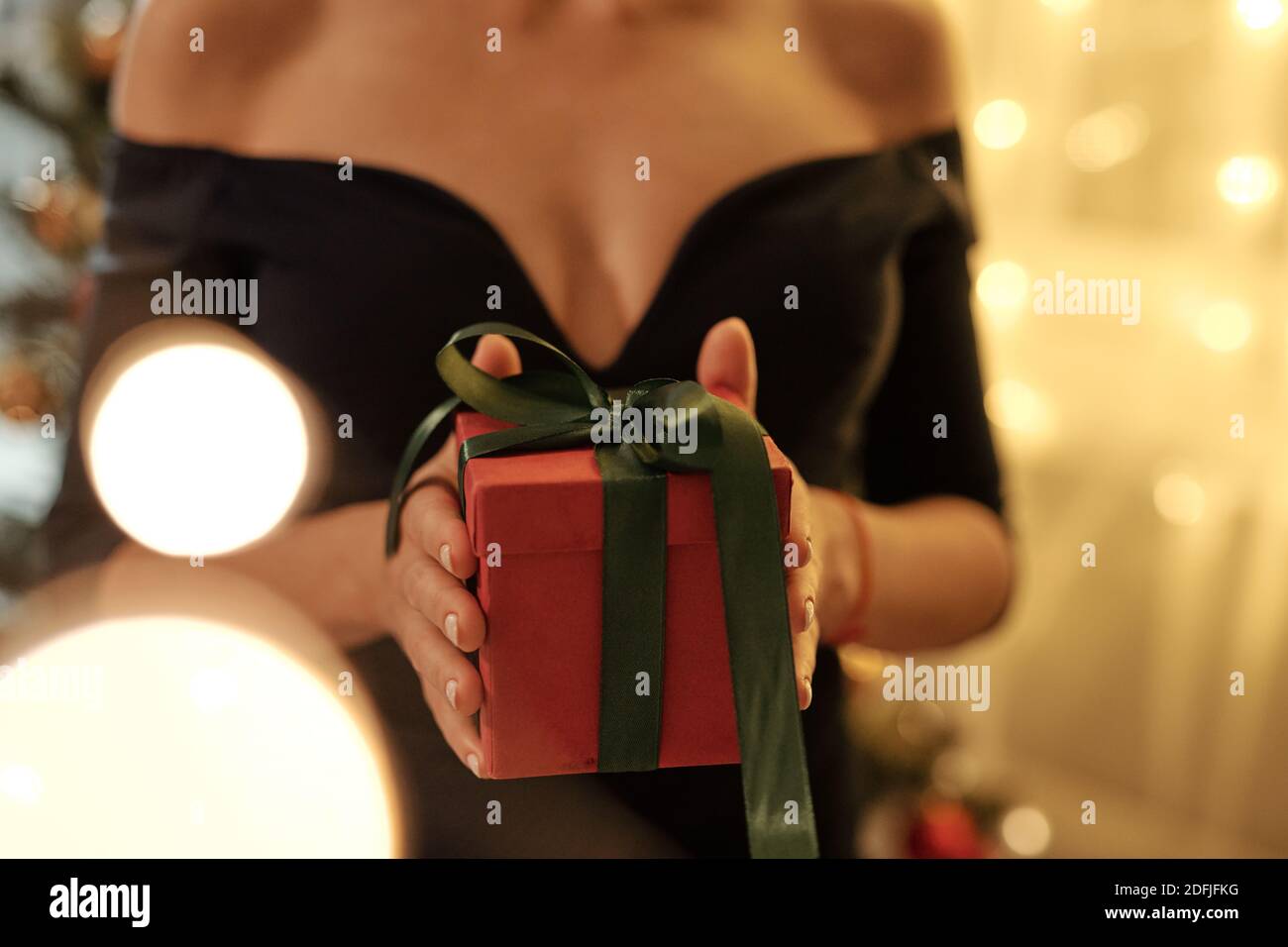 Hände von jungen eleganten weiblichen in schwarzen Abendkleid halten Rot verpackte geschenkbox gebunden mit dunkelgrünem Band im Stehen Vor der Kamera Stockfoto