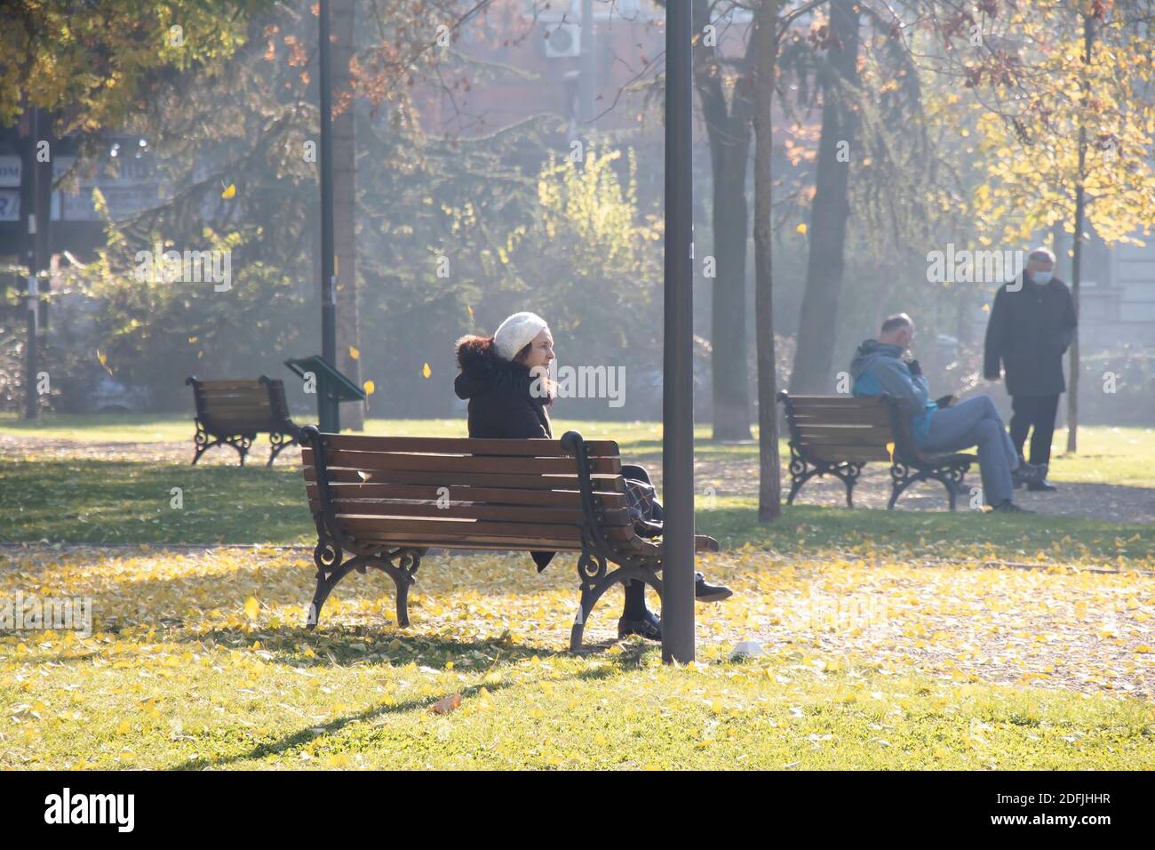 Belgrad, Serbien - 1. Dezember 2020: Eine reife Frau sitzt allein auf einer Parkbank, mit Menschen hinter sich, an einem sonnigen kalten Herbsttag Stockfoto