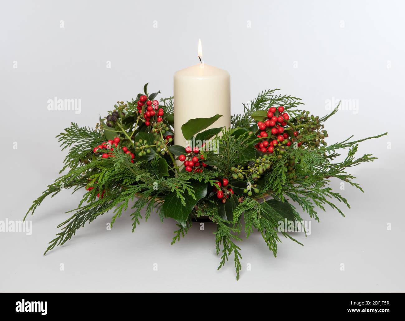 Isolierte, hausgemachte festliche Weihnachtsdekoration mit Holly Beeren, Fir, Efeu und einer brennenden Kerze vor einem schlichten Hintergrund. Stockfoto