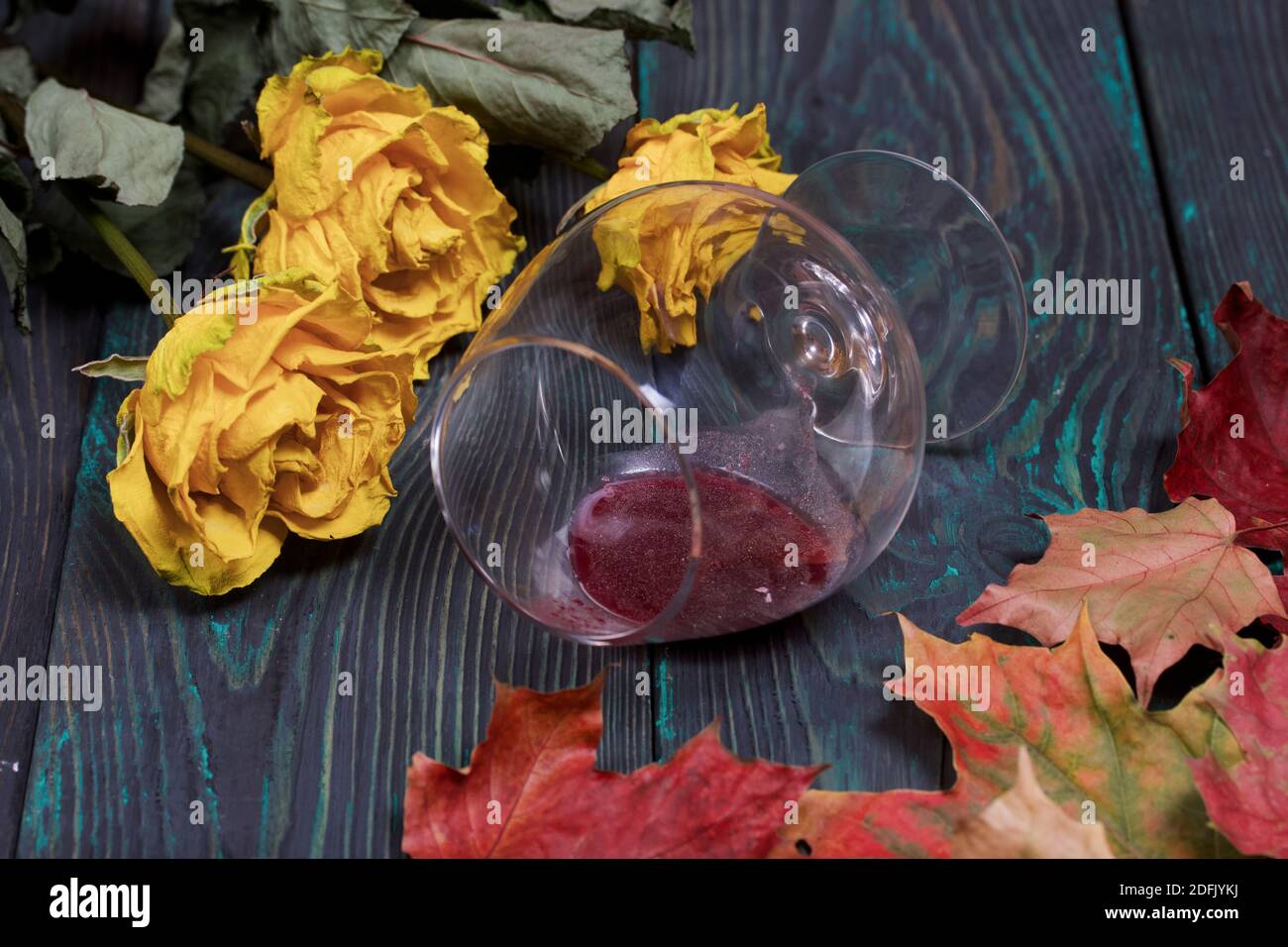 Ein Bouquet von getrockneten gelben Rosen und getrockneten Herbstblättern. Ein umgedrehtes Glas Sauerwein. Auf Kiefernbrettern. Stockfoto
