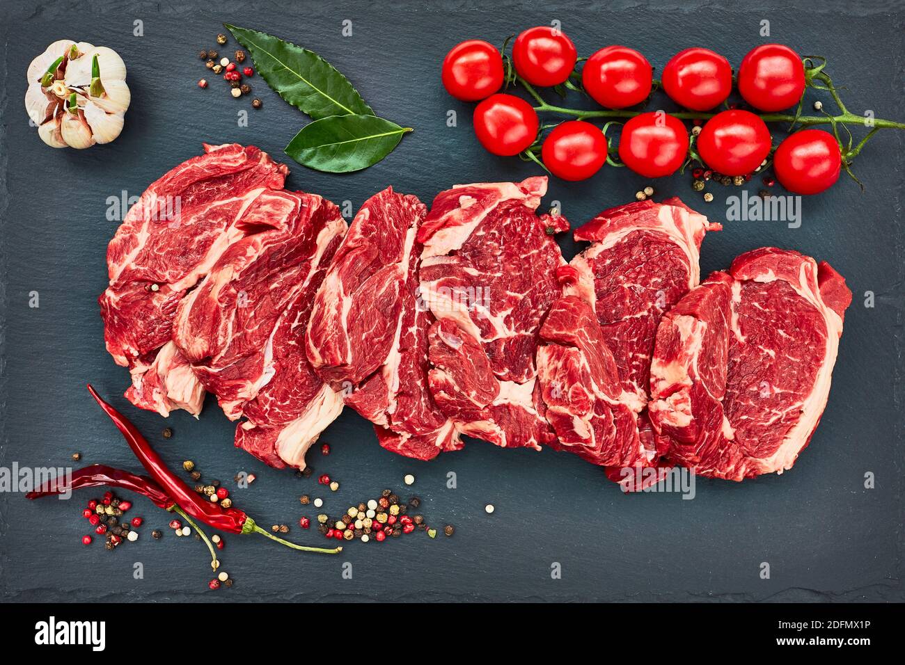 Frische rohe Rindersteaks mit Pfeffer und Tomaten auf schwarzem Schieferbrett. Platz kopieren, Draufsicht. Stockfoto