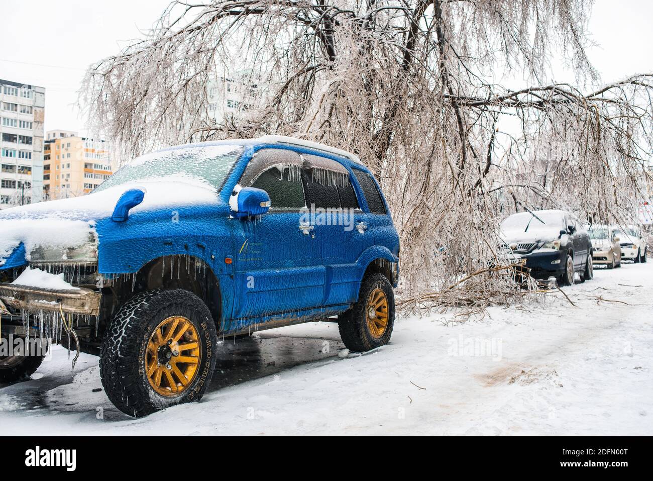 Blaues Auto bedeckt mit Eis und Eiszapfen nach kaltem Regen. Bäume, die mit Eis bedeckt sind und zu Boden gebogen sind. Stadt im Winter. Eissturmzyklon. Stockfoto