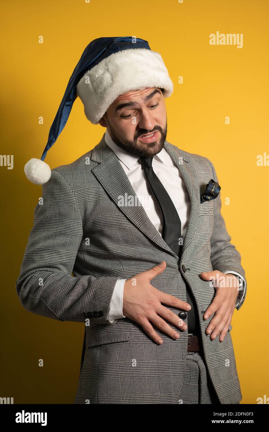 Overeat junge gut aussehende bärtige Mann berühren seinen Bauch trägt blauen Weihnachtsmann Hut und Anzug, Blick auf die Kamera isoliert auf gelbem Hintergrund. Positiv Stockfoto