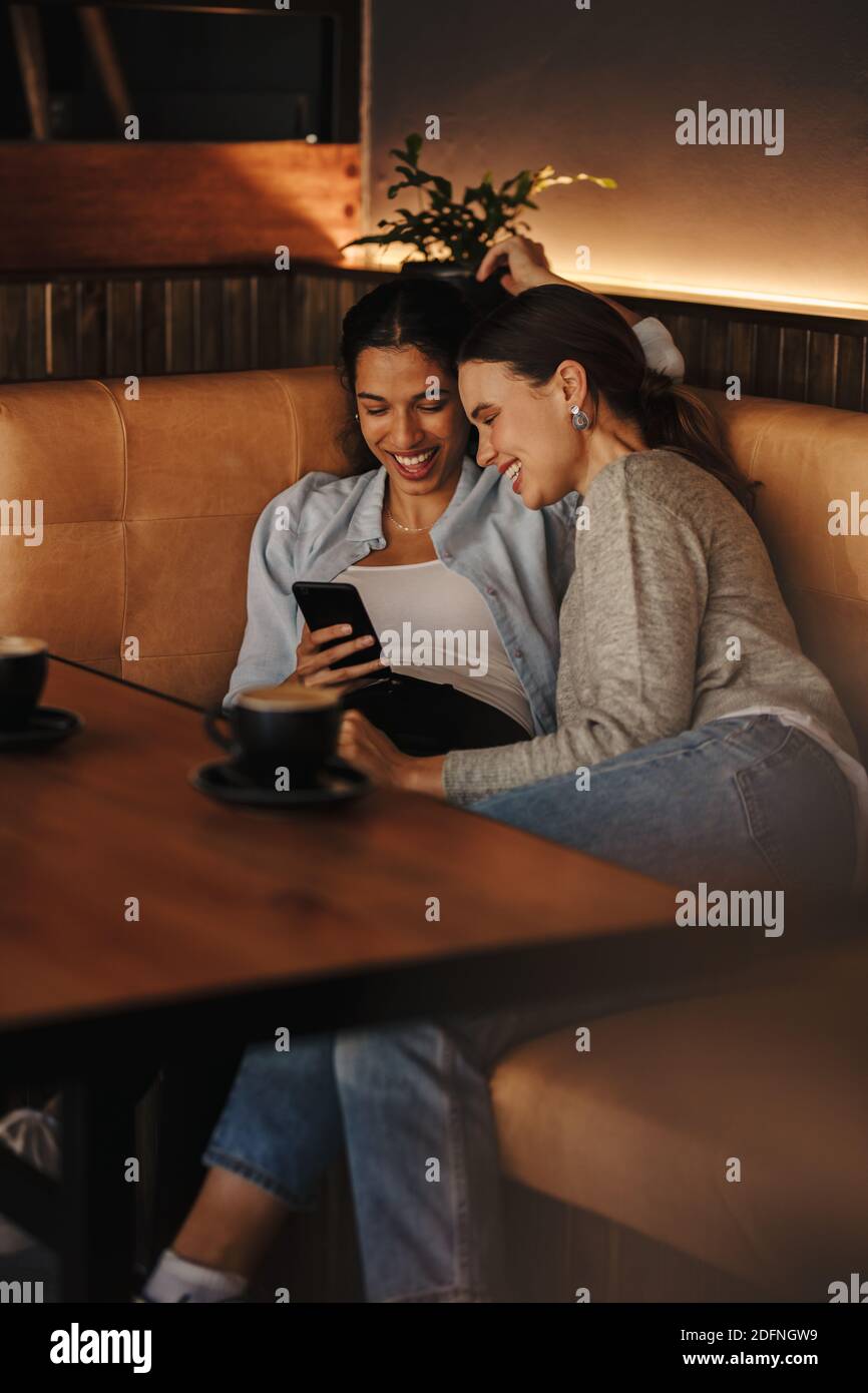Glückliche Freundinnen in einem Café an einem Wochenende. Zwei Frauen, die an einem Couchtisch sitzen und lächelnd auf das Smartphone schauen. Stockfoto