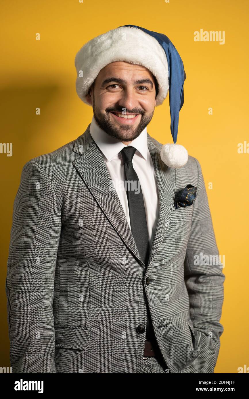 Junger schöner bärtiger lächelnder Weihnachtsmann mit blauem Weihnachtsmann Hut und Anzug, Blick auf die Kamera isoliert auf gelbem Hintergrund. Positive Emotionen Stockfoto