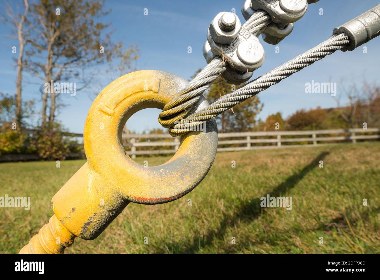 Gelber Spanndraht für Stahlseil. Zwei Stahlseile werden durch den Anker geschleift. Bäume und Zaun im Hintergrund. Anker ist im Gras. Stockfoto