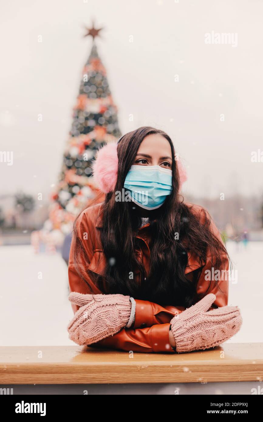 Glückliche junge schöne Frau in medizinischen Gesichtsmaske, pelzigen Ohrenschützer, rosa Fäustlinge auf Outdoor-Eislaufbahn. Fröhliche attraktive Mädchen in schützenden Mas Stockfoto