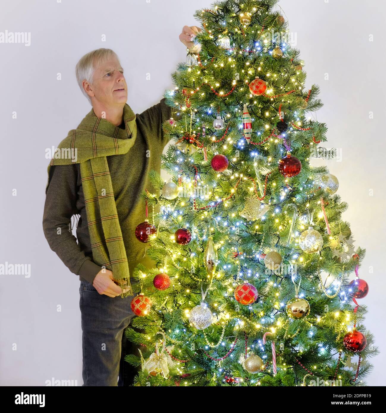 Ein reifer Mann mit grauen Haaren trägt grünen Pullover und Schal Platzierung einer Dekoration auf einem hell erleuchteten Weihnachtsbaum. Glaskugeln in Gold und Rot Stockfoto