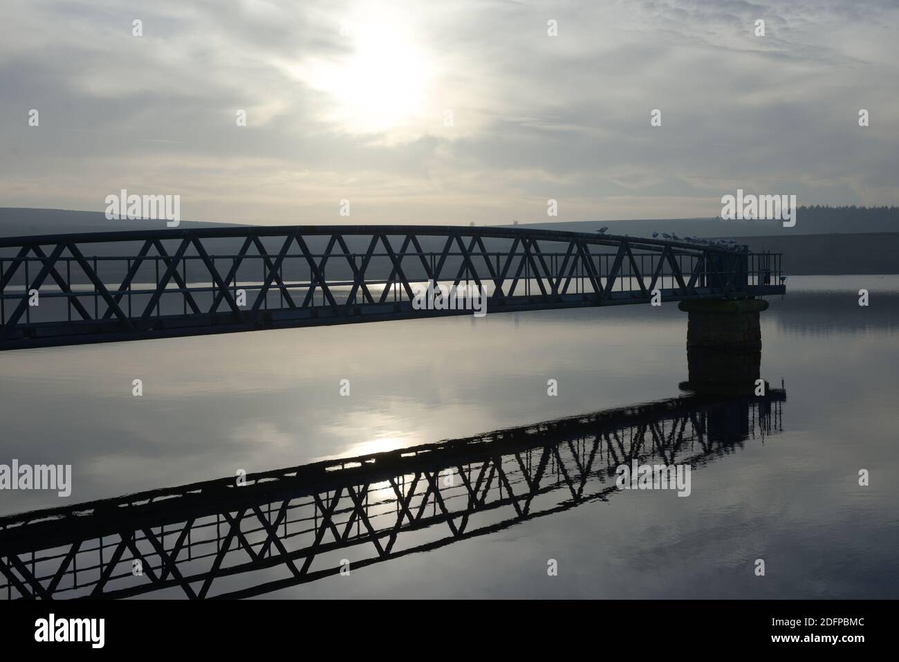 Vögel setzen sich auf einem Metallsteg mit ruhigem Wasser und abendlicher Sonneneinstrahlung zurück. Die luftige Atmosphäre steht im Kontrast zu den kühnen Stahlstreben. Stockfoto