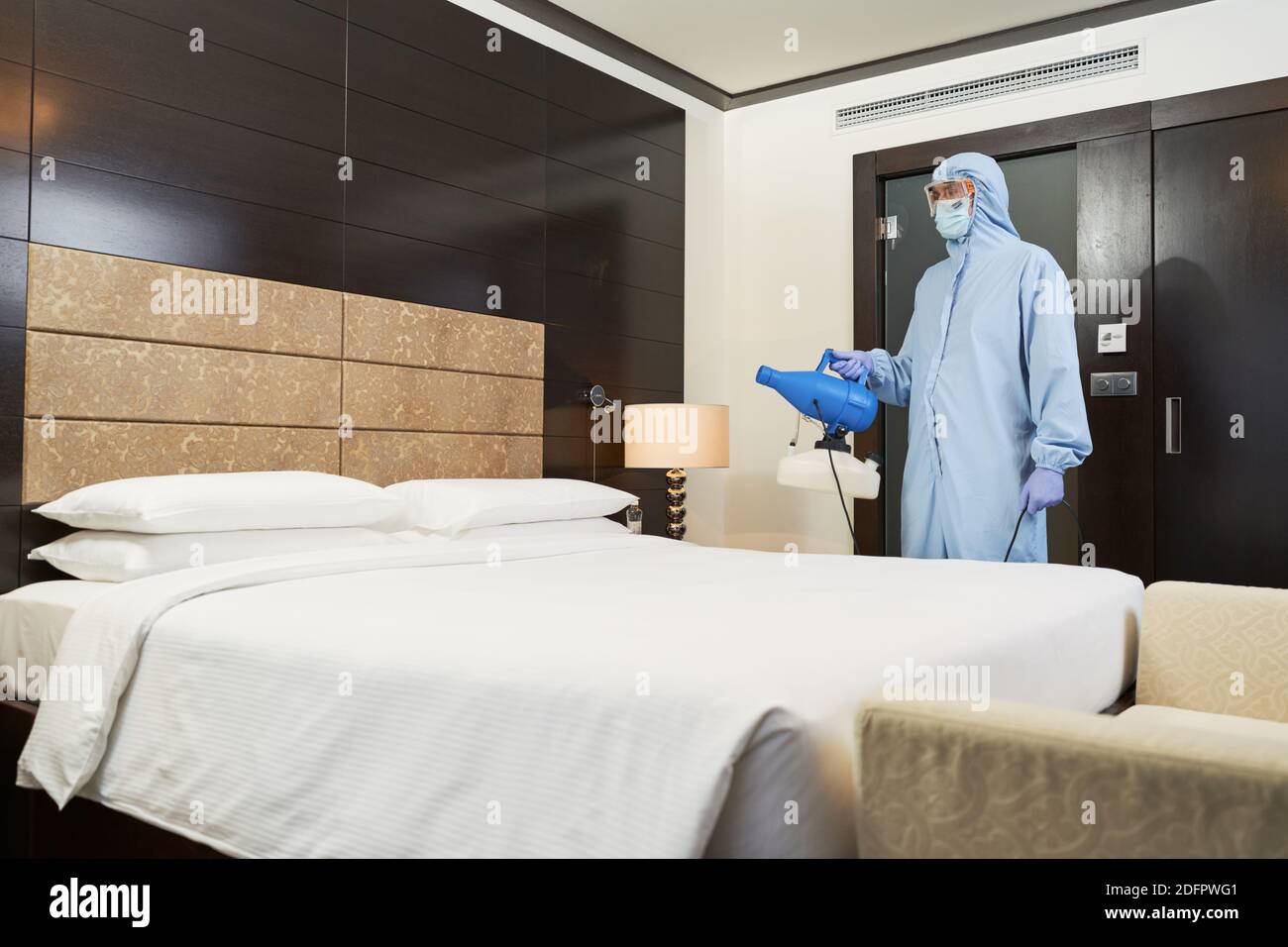 Männliche Desinfektionsmittel im Schutzanzug und Maske sprühen Desinfektionsmittel im Hotelzimmer. Coronavirus und Quarantänekonzept Stockfoto