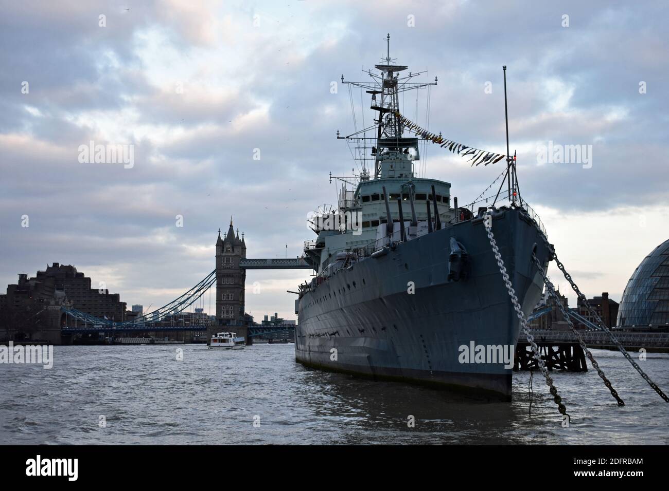 HMS Belfast liegt an der Themse. Hinter dem Kriegsschiff Museum ist das Rathaus und die victoria Tower Bridge an einem bewölkten Tag in London, Großbritannien. Stockfoto