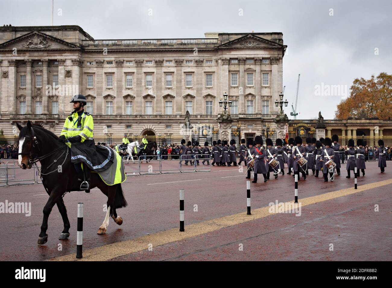 Ein Polizist auf einem Pferd führt die Soldaten in Bärenfellen und grauen langen Mänteln während der Wachwechselparade am Buckingham Palace in London. Stockfoto