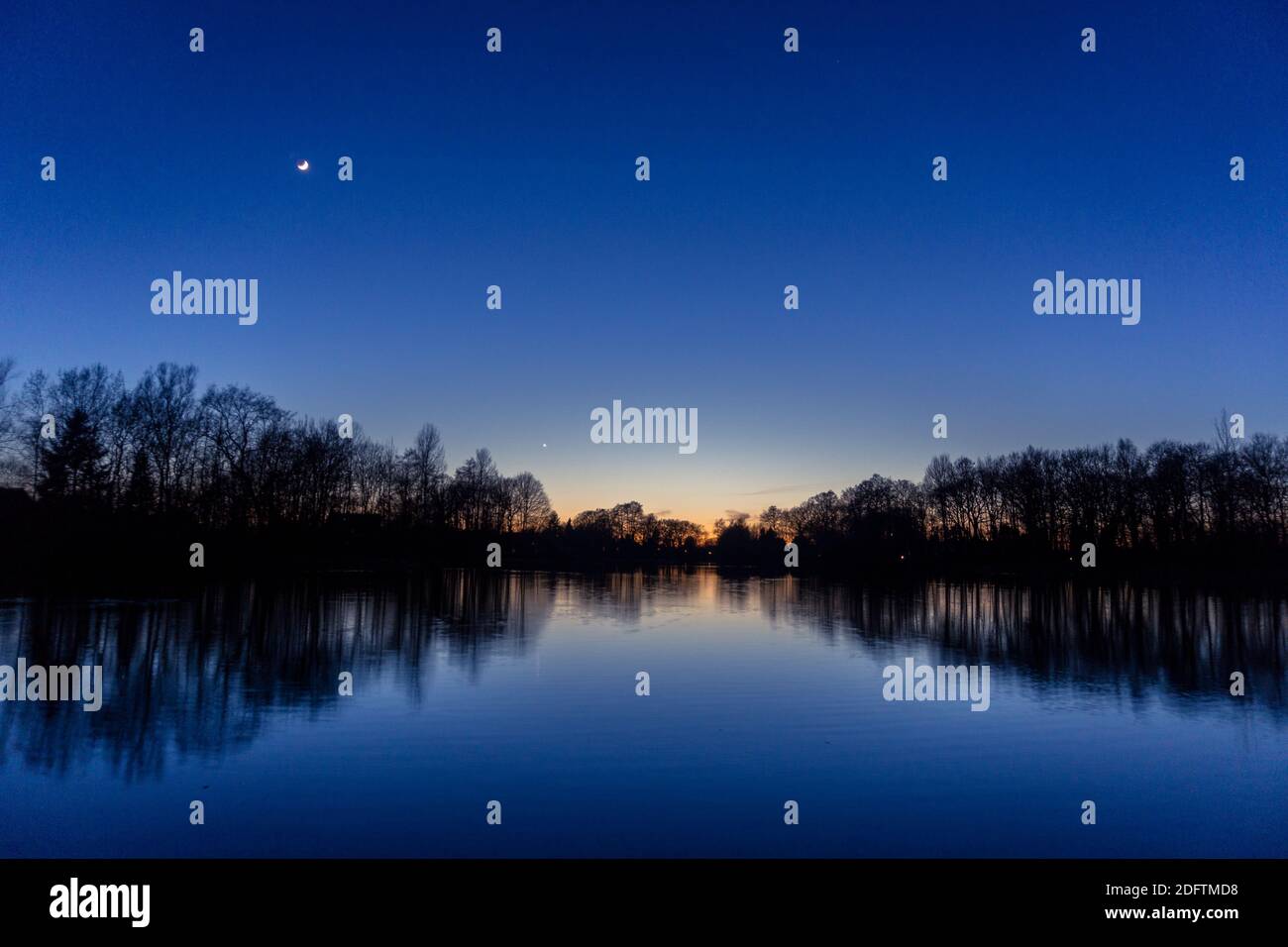 Klarer blauer Himmel nach Sonnenuntergang am See mit reflektierenden Silhouetten von Bäumen, venus und Halbmond Stockfoto