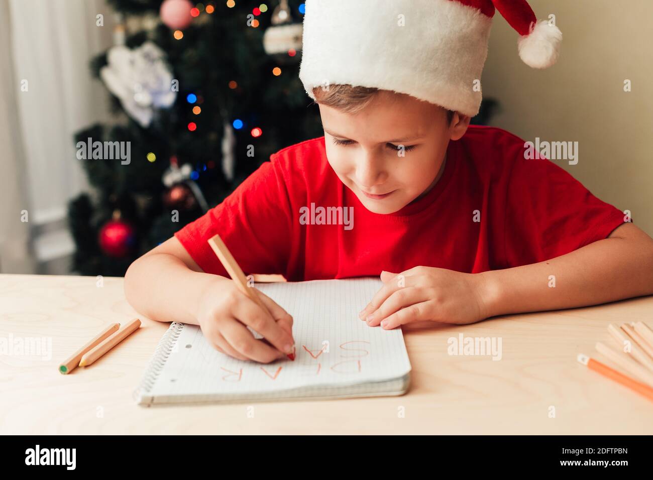 Lächelndes Kind Junge trägt roten Hut schreiben Brief an den Weihnachtsmann sitzen am Schreibtisch, Weihnachtsbaum auf dem Hintergrund, Haus innen Stockfoto