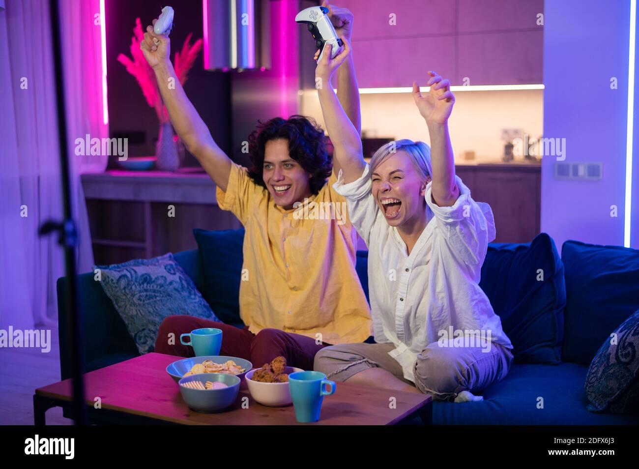 Lächelndes Paar hält Gamepads spielen Videospiel zu Hause. Low-Angle-Aufnahme junger Menschen, die während der Selbstisolation gegen Pandemie Zeit miteinander verbringen. Stockfoto