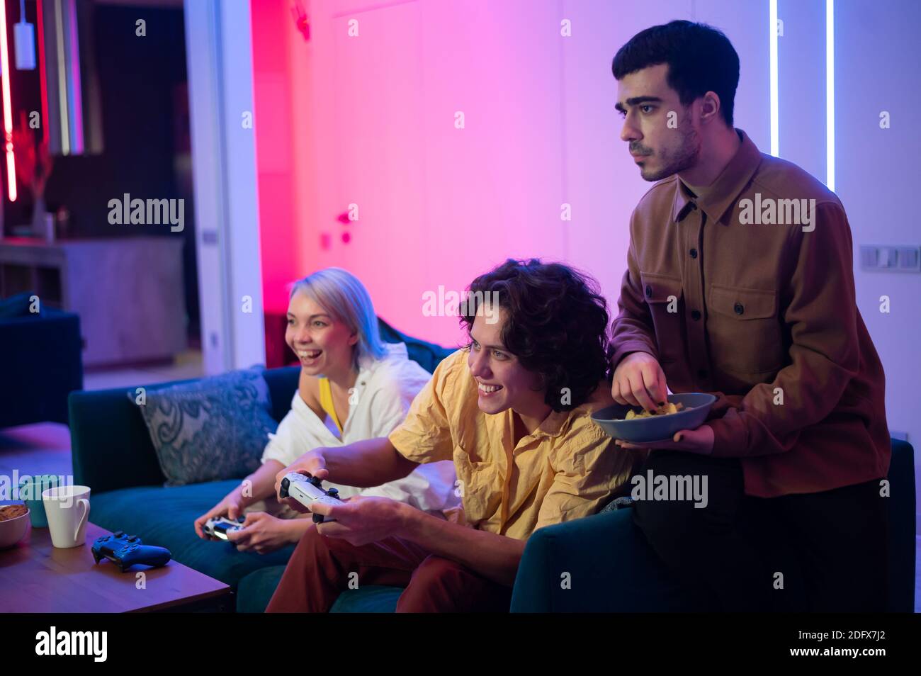 Zwei männliche Freunde aufgeregt, Videospiele zu Hause zu spielen, während ein Mädchen mit ihnen auf einer Couch und mit Handy-Smartphone sitzen. Stockfoto
