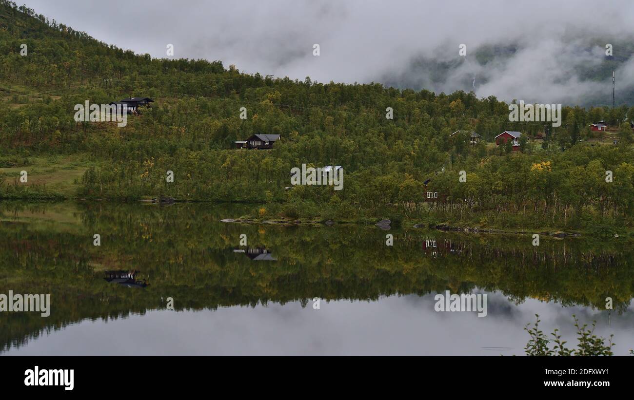 Traditionelle Holzhäuser umgeben von grünem Wald am Ufer des Lapphaugvatnet Sees im Norden Norwegens spiegeln sich im ruhigen Wasser am bewölkten Tag wider. Stockfoto