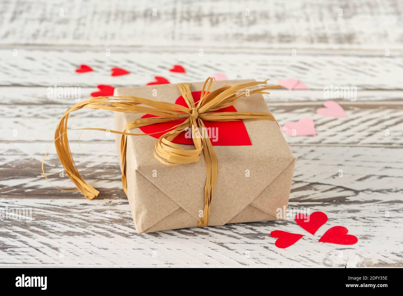 Bastelpapier Geschenkbox mit Kordelschleife und roten Herzen auf Holzhintergrund. Festliche Grußkarte zum Valentinstag und Geburtstag. Stockfoto