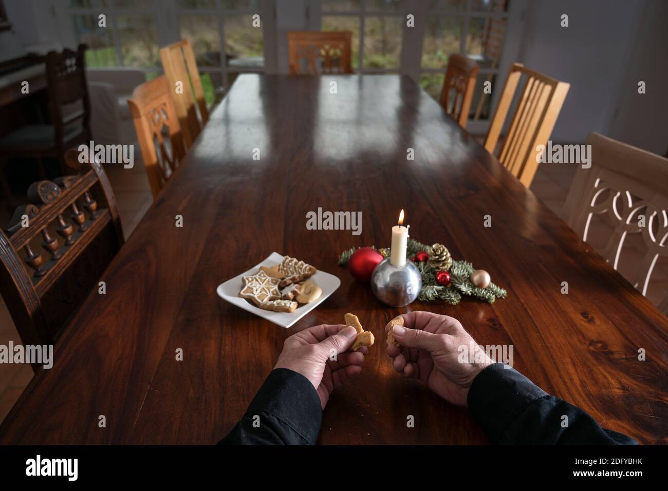 Hände eines älteren Mannes brechen ein Cookie an einem großen Familientisch mit Kerze, Weihnachtsdekoration und vielen leeren Stühlen, einsame Ferien während der Th Stockfoto