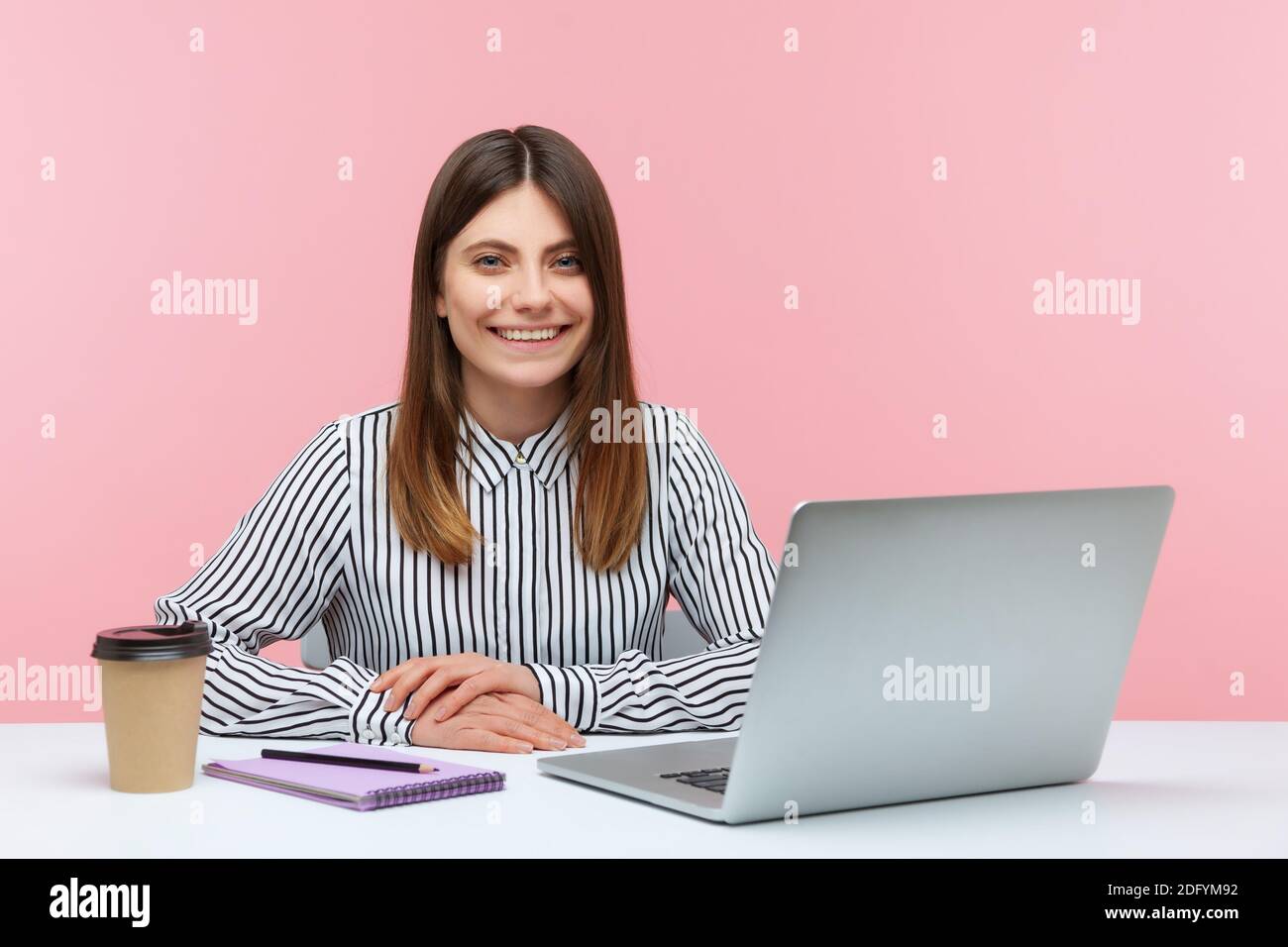 Happy fröhlich Büro Frau im gestreiften Hemd sitzenden Arbeitsplatz Blick auf die Kamera mit toothy Lächeln, Kaffee trinken und Notizen arbeiten auf Laptop Stockfoto