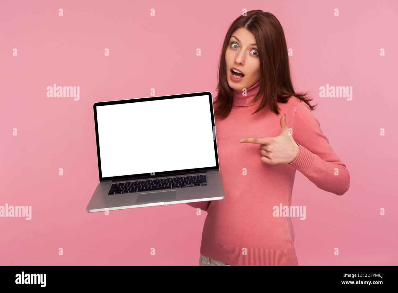 Glücklich überrascht Brünette Frau in rosa Pullover zeigt auf leeren Laptop-Bildschirm, zeigt leere Display für Web-Werbung. Innenaufnahme des Studios isoliert Stockfoto