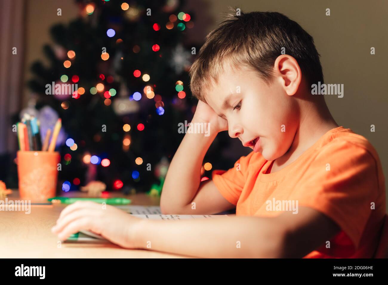 7 Jahre alter Junge, der am Schreibtisch sitzt und Buch liest. Geschmückter Weihnachtsbaum im Hintergrund Stockfoto