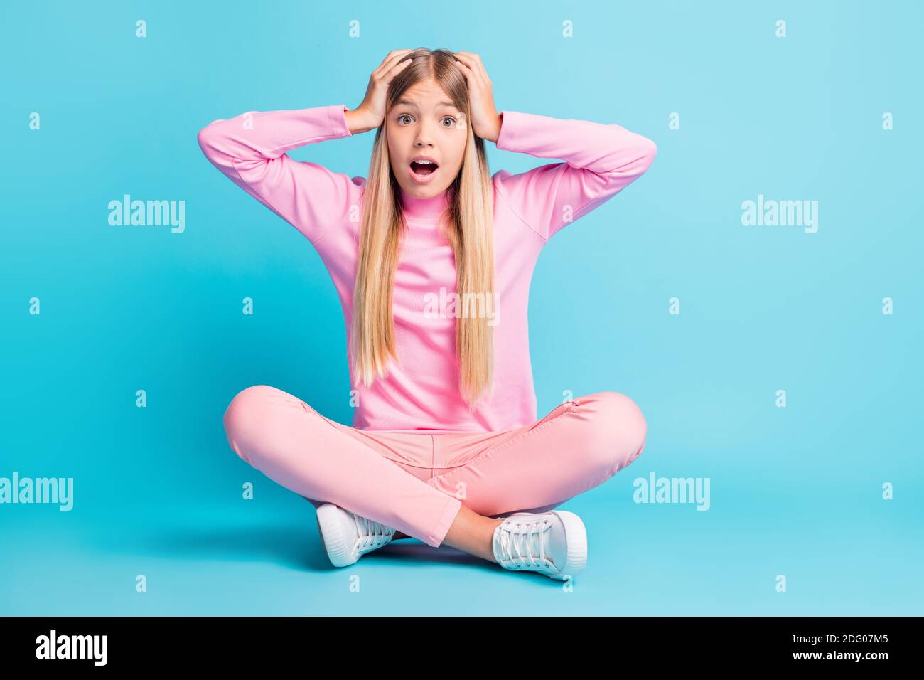 Foto von beeindruckt ziemlich blonde Teenager Mädchen sitzen Lotus offen Mund Arme Kopf Kleid rosa Hose Poloneck Schuhe isoliert auf blaugrün Hintergrund Stockfoto