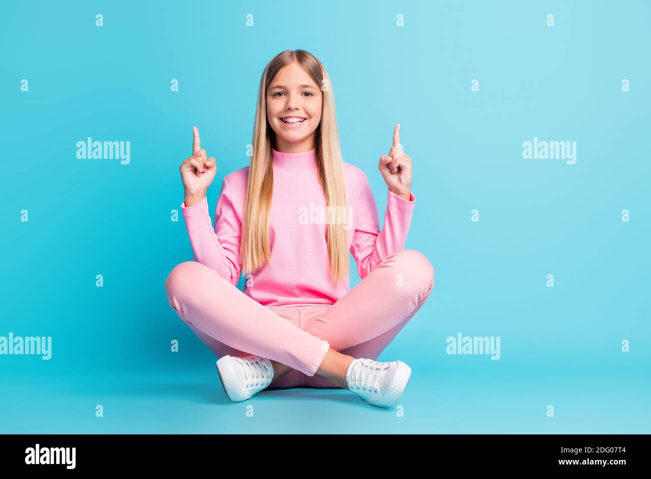 Foto von positiven ziemlich blonde Teenager-Mädchen sitzen Lotus Point Leerer Raum Kleid rosa Hose Poloneck Schuhe isoliert auf teal Hintergrund Stockfoto