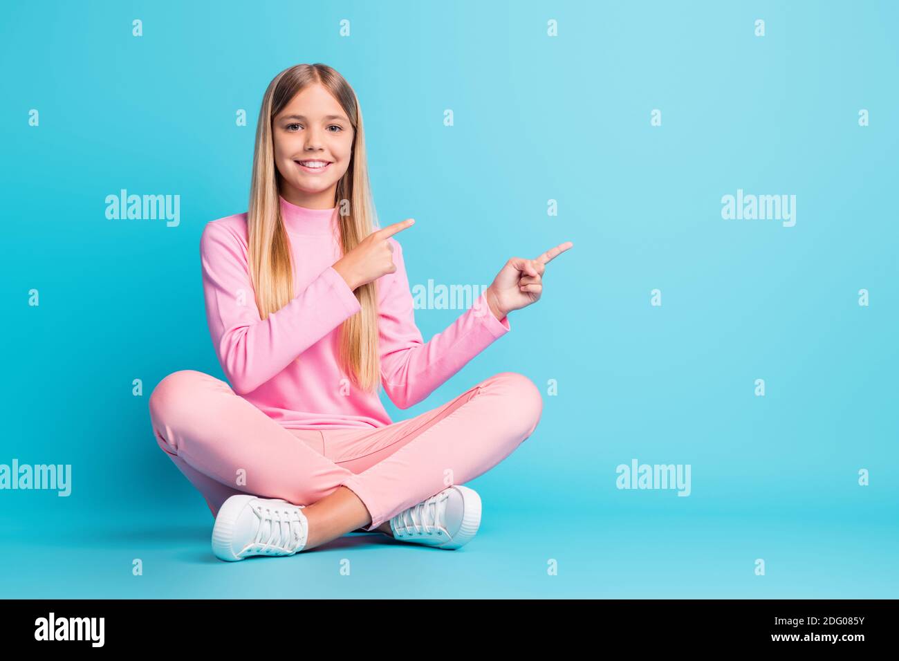 Foto von fröhlichen schönen blonden Teenager-Mädchen sitzen Lotus Point Leerer Raum Kleid rosa Hose Poloneck isoliert auf blauem Hintergrund Stockfoto