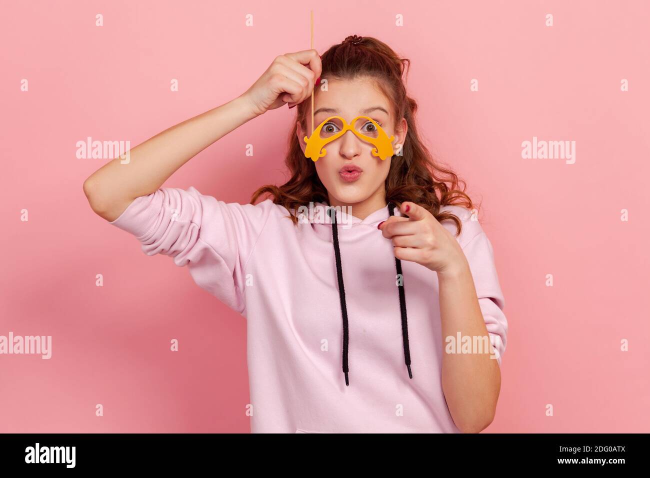 Portrait von fröhlichen Teenager-Mädchen mit Pout Lippen halten gedreht Papiergläser auf Stick und Zeigefinger, Spaß auf der Party, Karneval Accessoires. Stockfoto