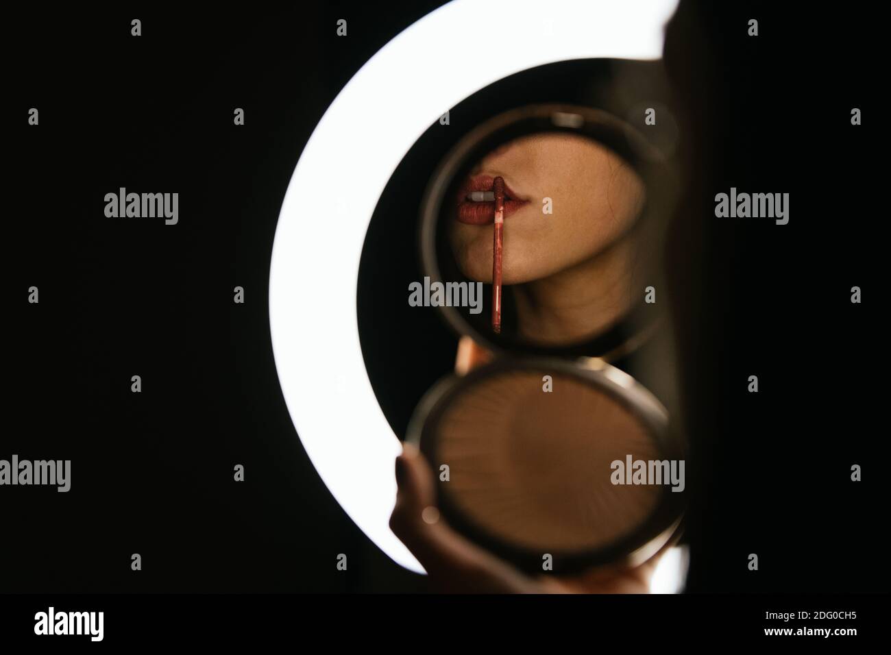 Schöne Frau malen ihre Lippen rot in einem runden Spiegel Beleuchtet mit einem Ring aus weißem Licht und einem schwarzen Hintergrund Stockfoto