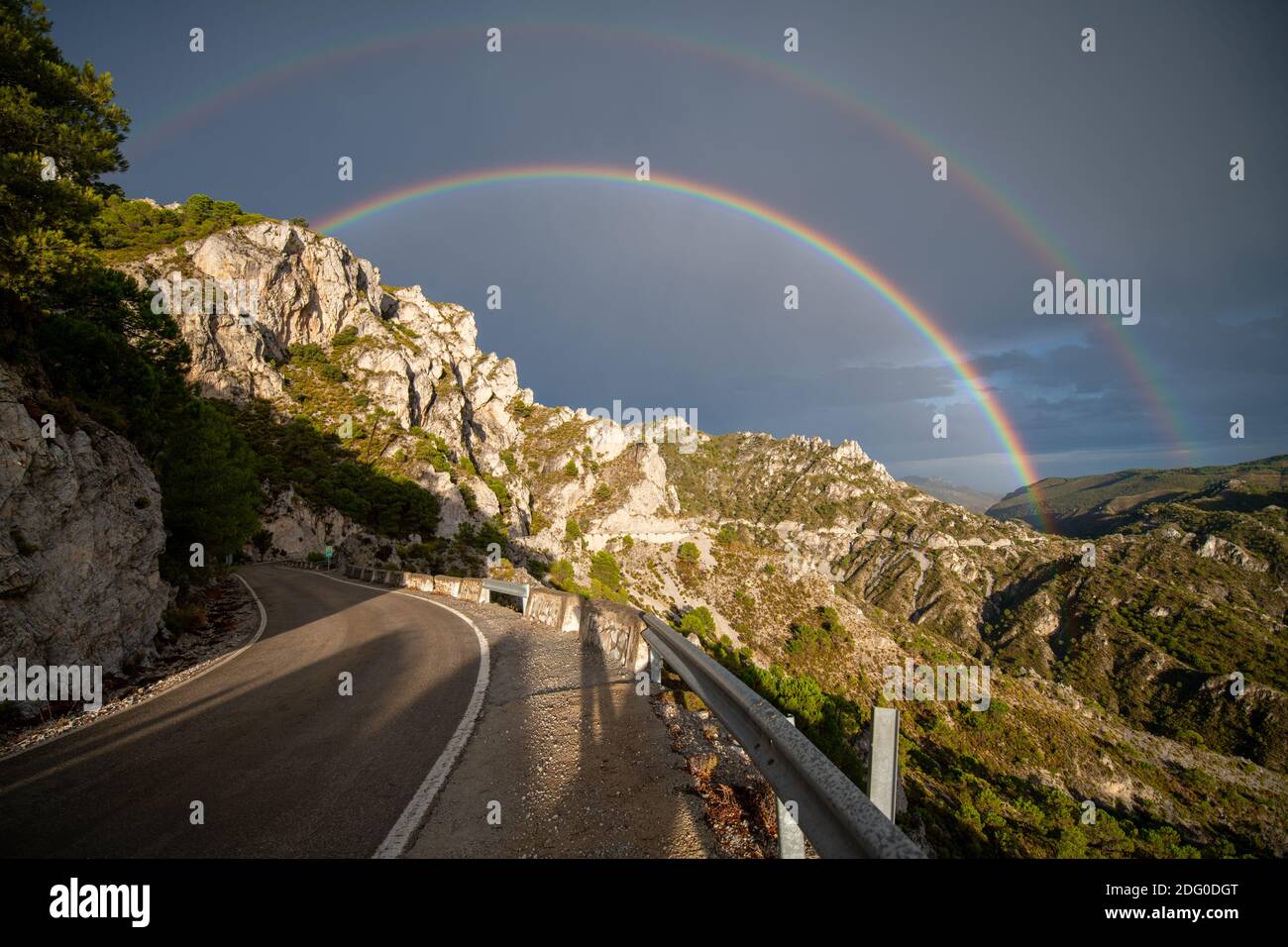 regenbogen über einer Bergstraße mit Klippen, hohen Gipfeln, grüner Vegetation und blau bewölktem Himmel Stockfoto