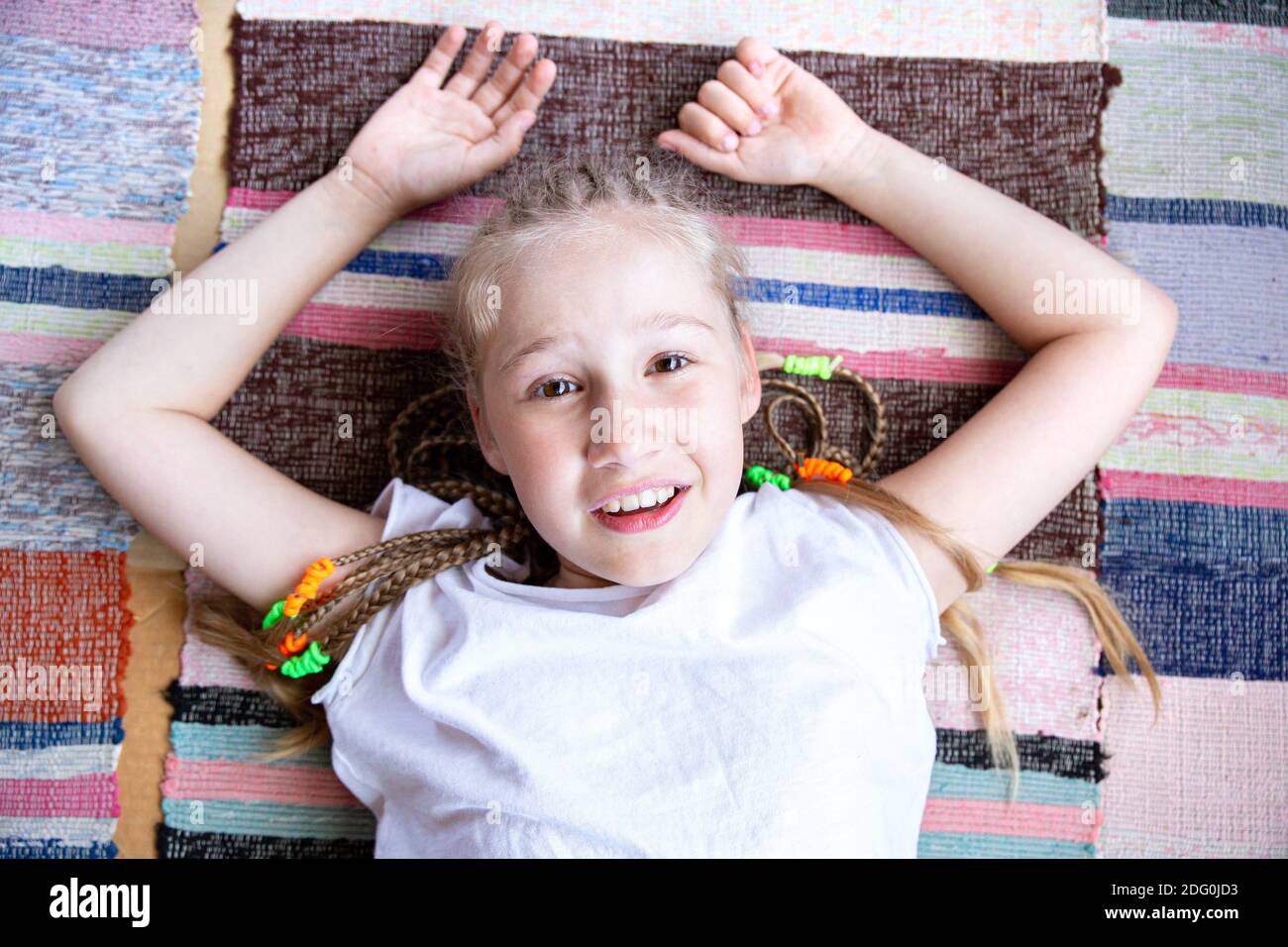 Porträt eines schönen lächelnden Mädchen mit Zöpfen Blick auf Kamera und lächelnd, auf einem gewebten Teppich mit den Armen nach oben liegend. Stockfoto