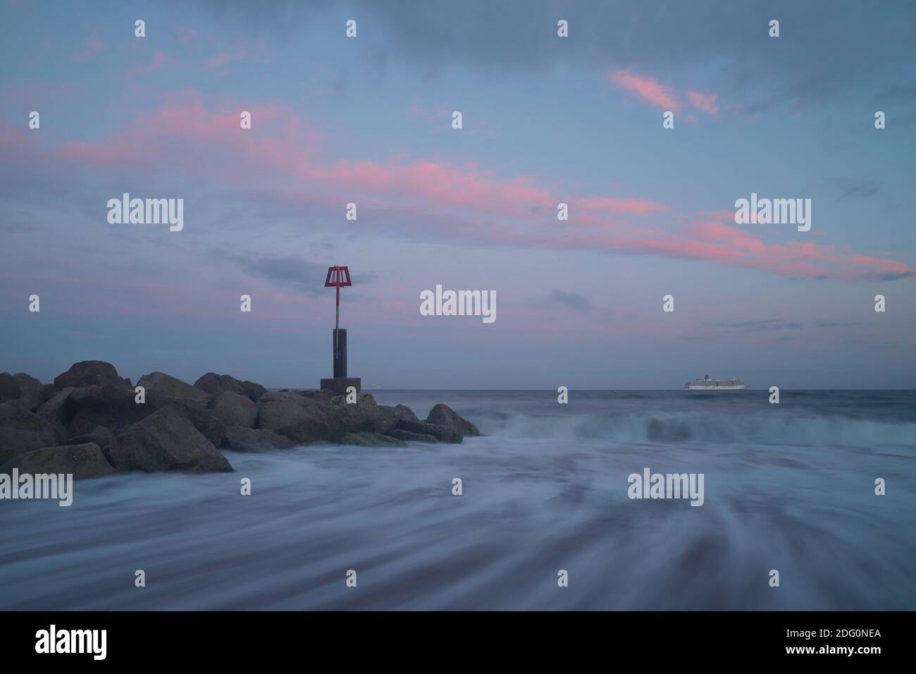 Bei Sonnenuntergang ziehen rosafarbene Wolken durch den Himmel und Wellen stürzen am Ufer um die Basis einer Groyne, während ein Kreuzfahrtschiff am Horizont sitzt. Stockfoto