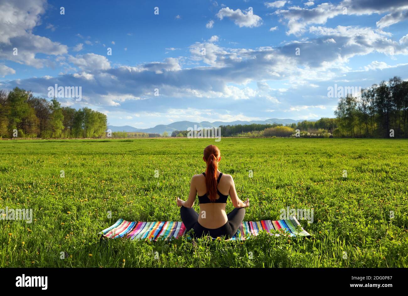 Auf dem Rasen sitzt das Mädchen in Lotushaltung auf der gestreiften Matte. Lifestyle Yoga Bewegung und Pose für ein gesundes Leben. Frühlingslandschaft. Landschaft mit Stockfoto