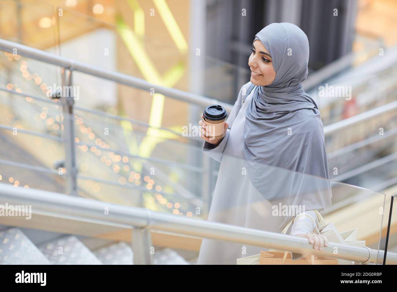 Porträt einer lächelnden Frau aus dem Nahen Osten, die Kaffeetasse hält, während sie im Einkaufszentrum die Treppe hinaufgeht und Platz kopiert Stockfoto