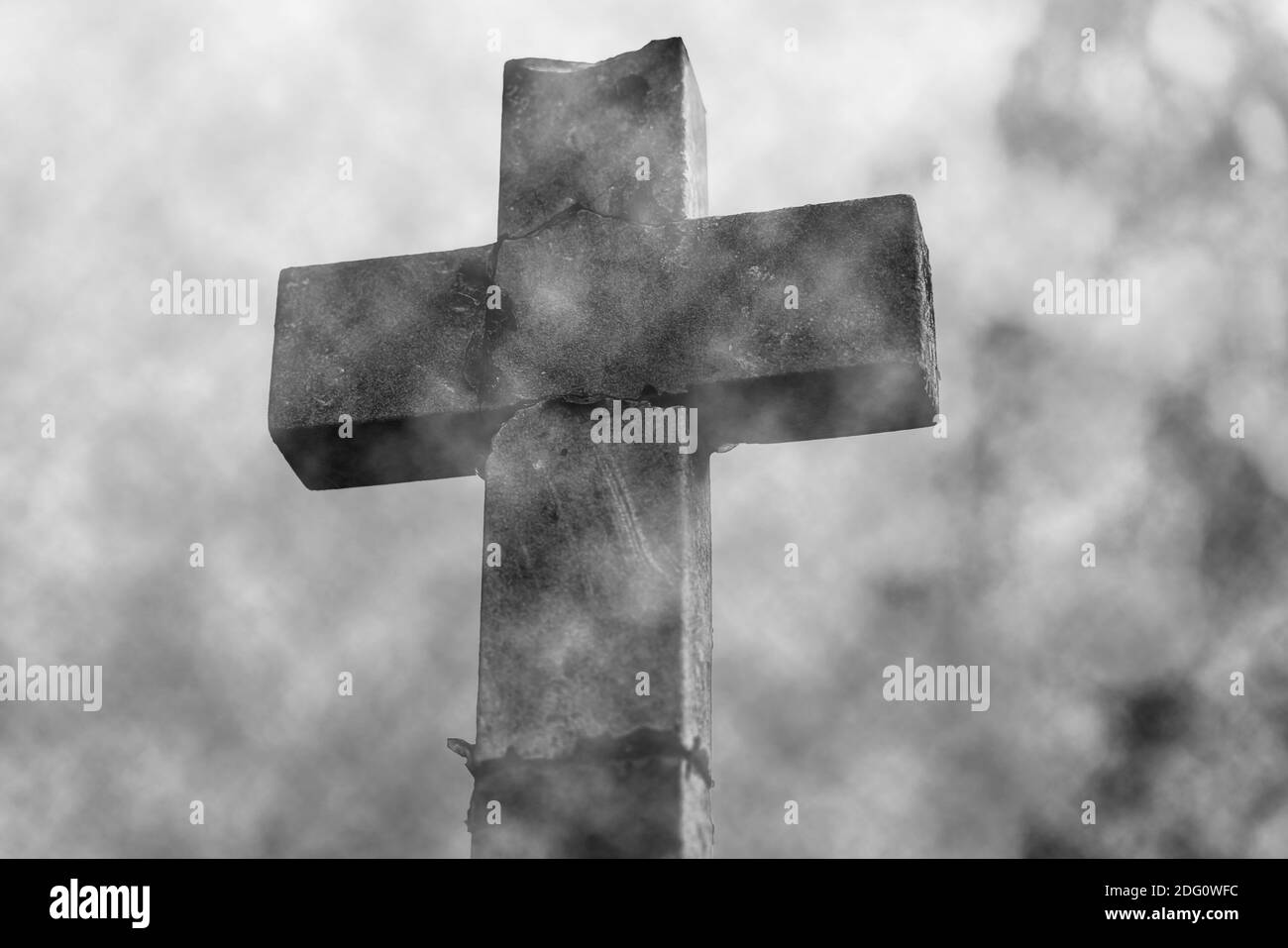 Alter verwitterter Steinkreuz-Grabstein auf einem Friedhof, der in schwerem weißem Nebel eingegossen ist. Schwarzweißbild mit verschwommenem Hintergrund. Stockfoto