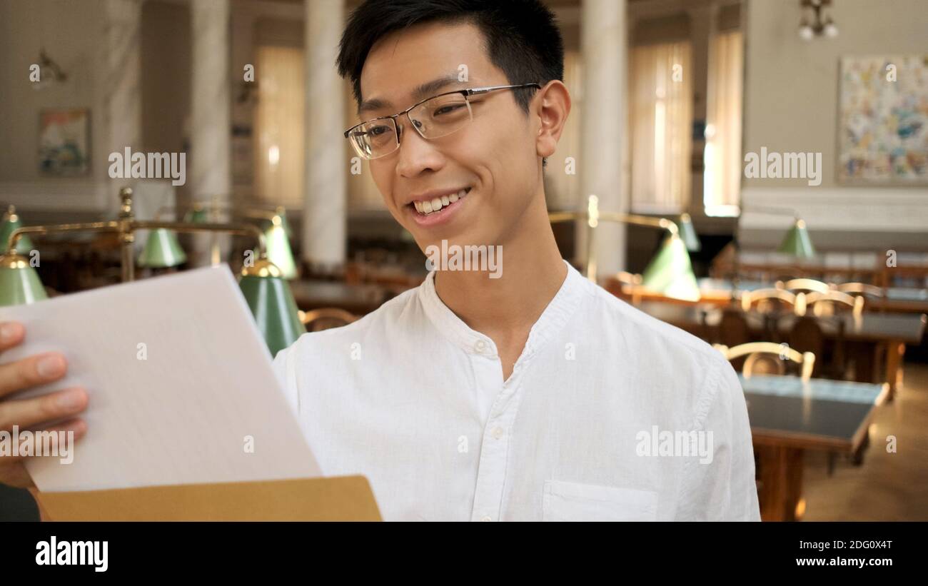 Portrait der asiatischen Student glücklich öffnen Umschlag mit Prüfungsergebnissen in der Universitätsbibliothek. Junge attraktive Kerl Öffnung Umschlag im Hochschule Stockfoto