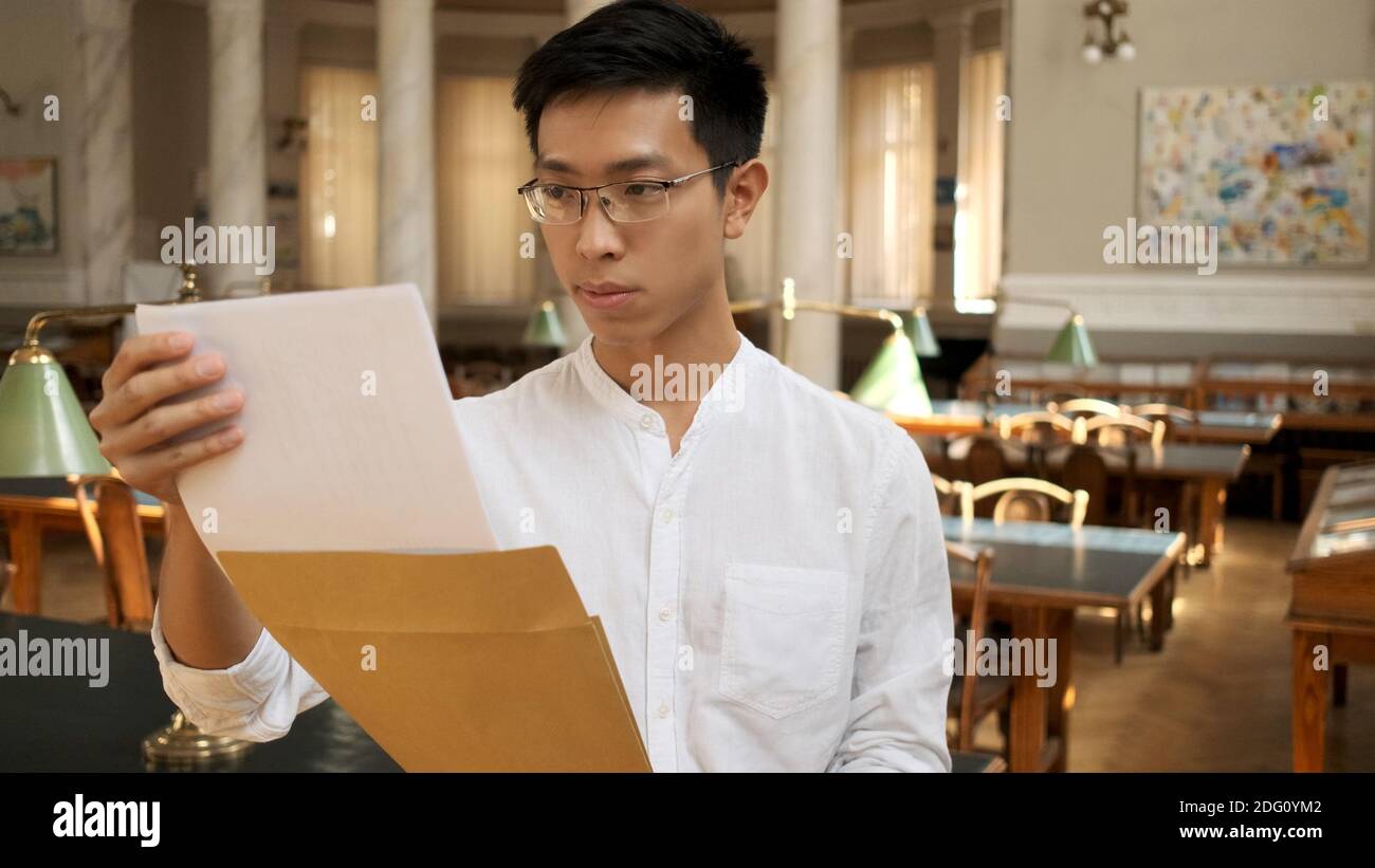 Asiatisch männlich Student intsently Öffnung Umschlag mit Prüfung Ergebnisse in Universitätsbibliothek Stockfoto