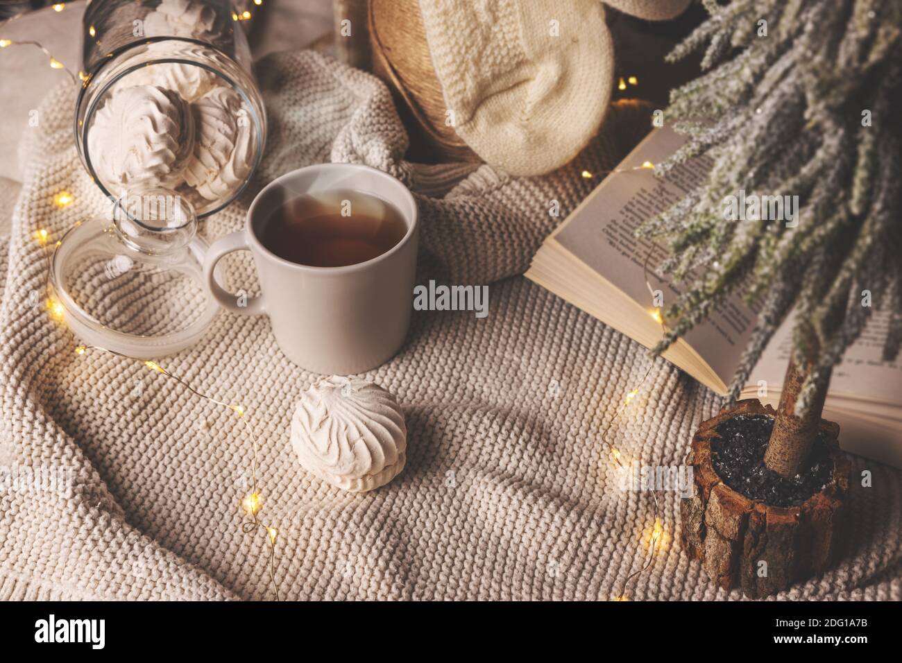 Warmes gemütliches Zuhause zu weihnachten - Tasse heißen Tee, Zephyren und Buch mit Lichtern auf Strickdecke Stockfoto