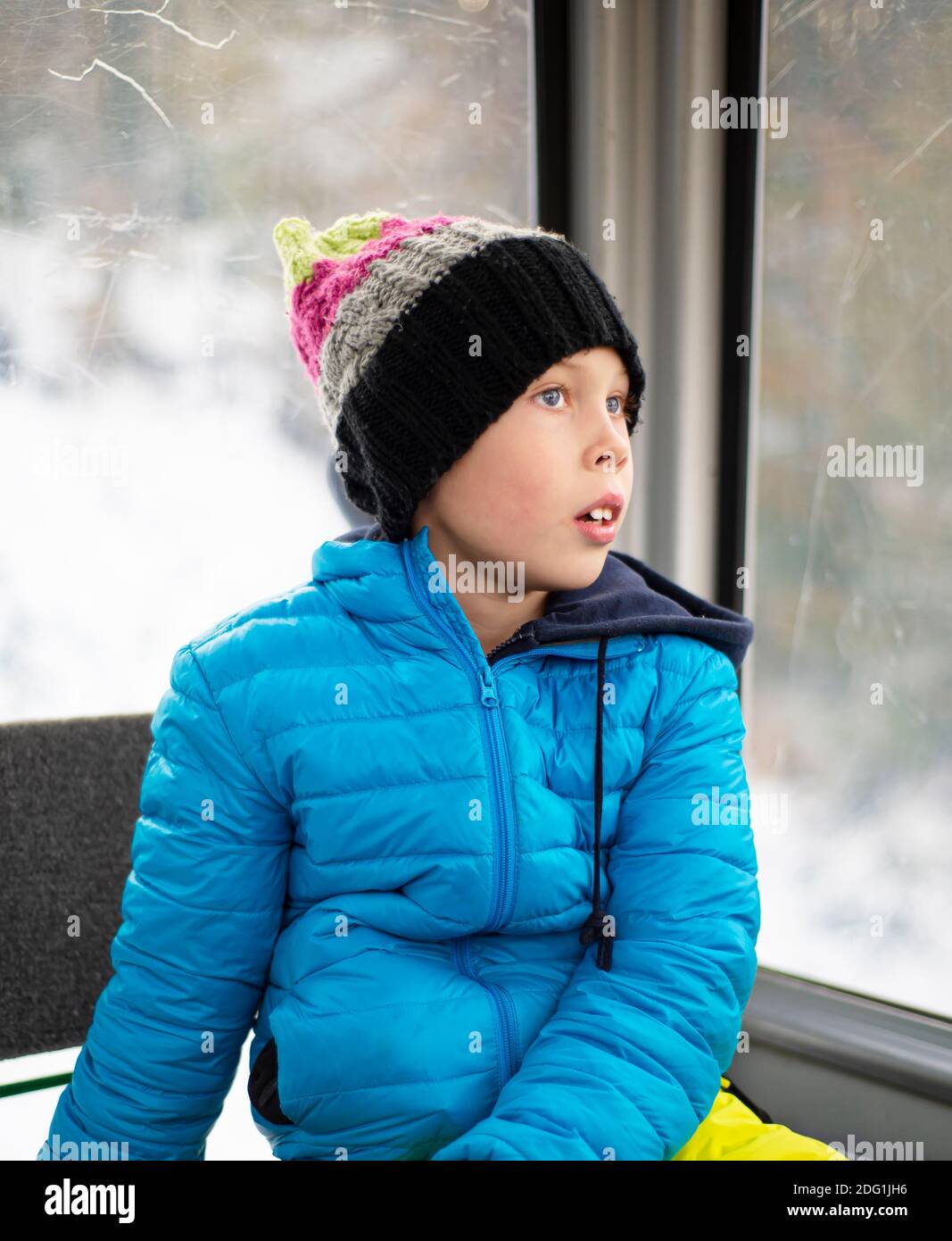 Ein Junge in einer blauen Jacke und einem gestrickten Hut sitzt in einer Kabine am Winterfenster und träumt. Seine blauen Augen blicken in die Ferne. Nachdenklicher Blick. Stockfoto