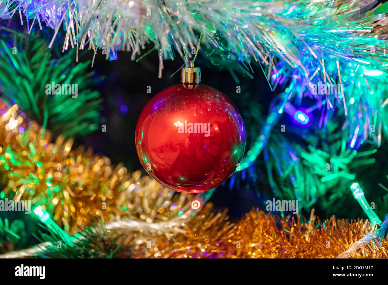 Dekorierter Weihnachtsbaum Nahaufnahme. Rote Kugel in der Mitte, beleuchtete Girlande mit blauen, orangefarbenen, grünen Lichtern. Neue Jahre, Winter Urlaub Licht de Stockfoto