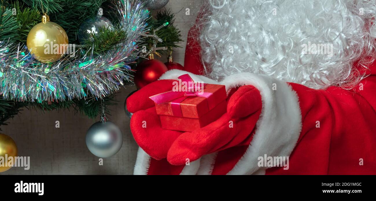 Nahaufnahme des Weihnachtsmannes mit einer roten Geschenkbox in beiden Händen. Weihnachtsbaum mit roten, weißen und goldenen Kugeln, weiße Lametta neben ihm. Neu Ja Stockfoto